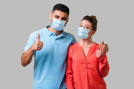 Portrait of positive young couple with surgical medical mask standing together, hugging as friends,...