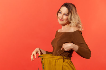 Portrait of beautiful young adult woman have willpower showing slim waist in big trouses, successful weight loss, diet conception. Indoor studio shot isolated on red backgroundの写真素材