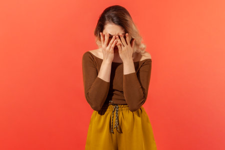 Oh no, it can't be! Portrait of sad young woman standing, who dramatic covered face with her palms, feeling trouble, pain and sorrow. Depressed concept. Indoor studio shot isolated on red backgroundの写真素材