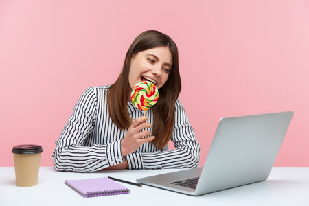 Funny playful woman licking and biting round colored lollipop on stick talking on video call at laptop sitting at workplace, having fun. Indoor studio shot isolated on pink backgroundの写真素材
