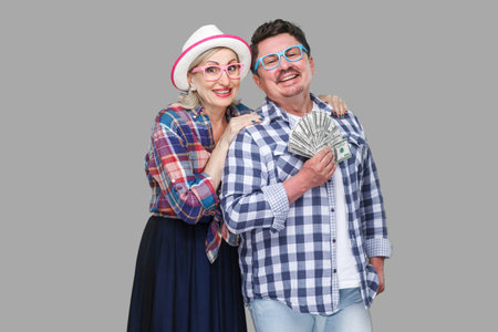 Happy wealthy family, adult man and woman in casual checkered shirt standing pickaback together, holding fan of dollar, toothy smile, looking at camera. Indoor,isolated, studio shot, gray backgroundの写真素材