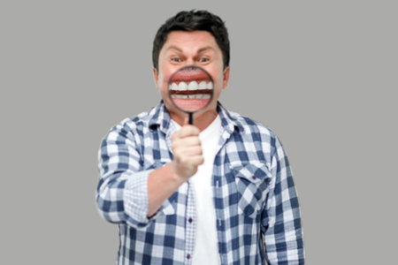 smile zoom or tooth care concept. Portrait of middle aged man in casual checkered shirt standing, holding magnifying glass on his teeth, looking at camera. studio shot, isolated on gray background.の写真素材