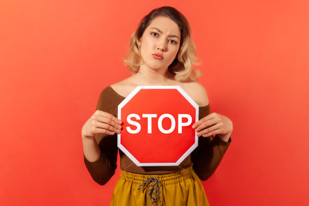 Serious woman holding and showing red stop sign, concerned about women rights, demanding equality. Indoor studio shot isolated on orange backgroundの写真素材