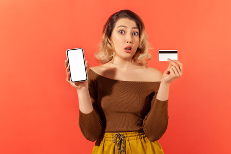 Shocked woman showing and holding empty screen smartphone and credit card, surprised with online crediting, internet banking. Indoor studio shot isolated on orange backgroundの写真素材