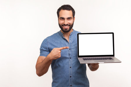 Cheerful positive man pointing finger showing empty display of laptop, blogger presenting freespace, advertising area. Indoor studio shot isolated on white backgroundの写真素材