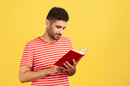 Concentrated bearded man holding book and smiling, enjoying reading, standing absorbed with exciting plot, checking schedule in notepad. Indoor studio shot isolated on yellow backgroundの写真素材