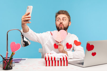 Funny bearded man office worker making selfie pressing lips in kiss holding paper pink heart, sitting at workplace all covered with heart shaped stickers. Indoor studio shot isolated on blue backgroundの写真素材