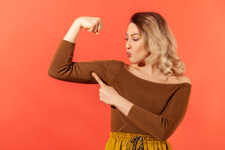 Strong independent woman with blond hair showing her arm muscle and point it with finger, proud of her strength and leadership skills, emancipation. Indoor studio shot isolated on red backgroundの写真素材