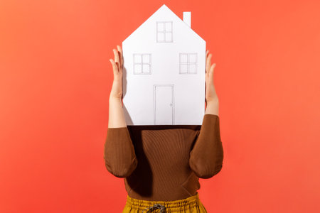 Woman hiding her face behind paper house with drawn windows and door, government housing program, real estate agency. Indoor studio shot isolated on orange backgroundの写真素材
