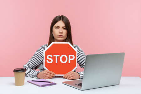 Serious woman office worker holding stop symbol, warning with red traffic sign and looking angrily,...