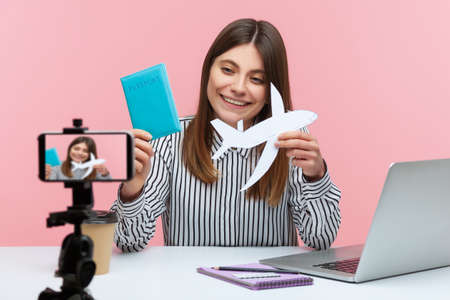 Positive woman blogger showing passport and paper plane at smartphone camera, bragging about tour, going to vacations. Indoor studio shot isolated on pink backgroundの写真素材
