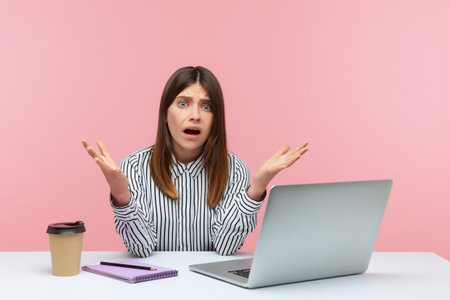 How could you? Shocked annoyed woman office worker raising hands in indignant gesture, asking why and looking angrily, sitting at workplace with laptop. Indoor studio shot isolated on pink backgroundの写真素材