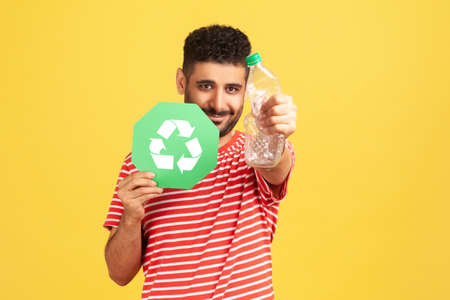 Pleased smiling bearded man in red striped t-shirt holding out hand with plastic bottle showing...