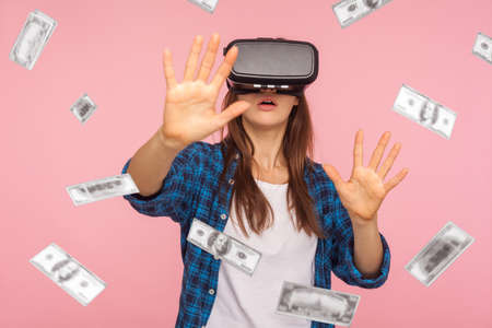 Portrait of shocked woman wearing virtual reality glasses, stretching hands forward to get money, dollar rain falling, illusion of rich millionaire, playing vr game, indoor isolated on pink backgroundの写真素材