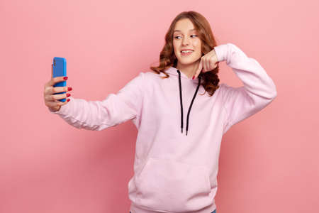 Portrait of joyful teen girl in hoodie pointing down while communicating by video call or streaming, gesturing to subscribe, taking selfie on smartphone. Indoor studio shot isolated on pink backgroundの写真素材