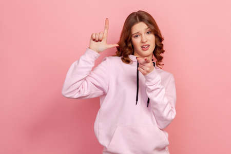 Portrait of bossy curly-haired teen expressing disrespect, showing L finger sign to camera, loser or lame gesture, accusing for failure. Indoor studio shot isolated on pink backgroundの写真素材