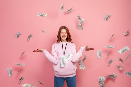 Full length portrait of cheerful young female in hoodie throwing dollar banknotes and sincerely smiling, lottery winner. Indoor studio shot isolated on pink backgroundの写真素材