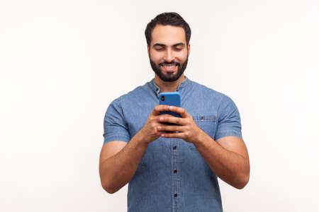 Smiling positive bearded man in blue shirt looking at smartphone display, surfing internet, betting, doing shopping online. Indoor studio shot isolated on white backgroundの写真素材