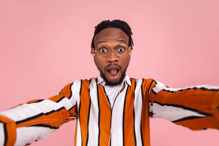 Surprised shocked african man blogger with beard and dreadlocks in striped shirt looking at selfie camera with astonishment, unbelievable news. Indoor studio shot isolated on pink backgroundの写真素材