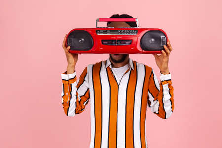 Funny afro-american man with beard and dreadlocks in bright striped shirt hiding his face behind red vintage audio record player, selling junk. Indoor studio shot isolated on pink backgroundの写真素材
