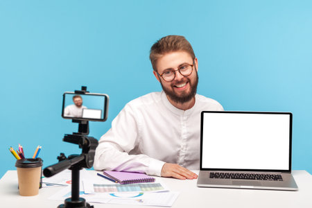 Positive smiling man office worker demonstrating laptop with white screen on smartphone camera,...
