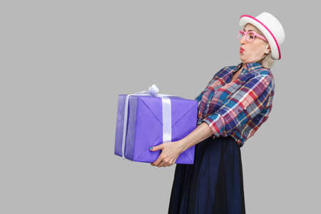 Side view portrait of cheerful wondering modern grandma in white hat and checkered shirt standing and trying to hold giant big heavy gift box and greeting you. Studio shot, gray background, isolatedの写真素材
