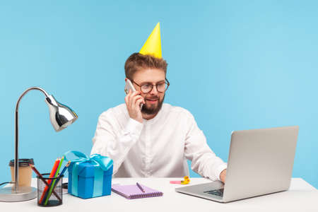 Busy birthday man with party hat on head receiving gifts and greetings talking phone, working on laptop, sitting at workplace. Indoor studio shot isolated on blue backgroundの写真素材