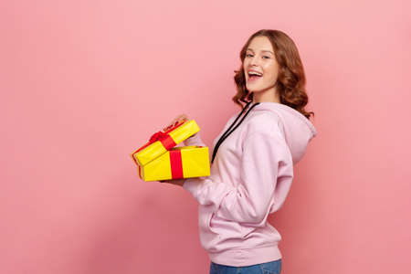 Profile of joyous curly haired teenage girl in hoodie standing with opened gift box, looking at...
