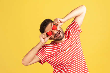 Funny positive man with beard in striped t-shirt having fun holding little toy hearts near eyes and...