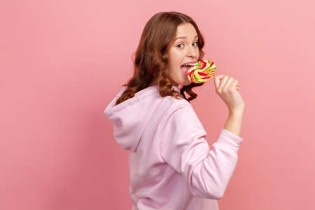 Side view portrait of funny curly haired teenage girl in hoodie licking round colorful lollipop...