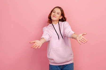 Portrait of kind young female with curly hair outstretching hands as if giving for free, offering to embrace, inviting with hospitable expression. Indoor studio shot isolated on pink backgroundの写真素材