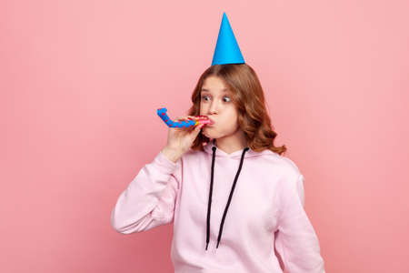 Portrait of funny curly haired teenage girl in hoodie with birthday hat blowing party pipe,...