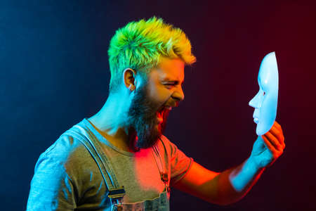 Profile of young adult attractive hipster man screaming angrily, holding white mask in hands, hating his hidden personality, expressing negative, hatred. Colorful neon light, indoor studio shot.の写真素材