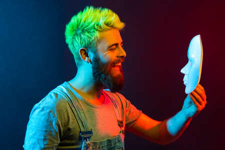 Happy bearded hipster man holding and looking at white mask with toothy smile, being satisfied of...