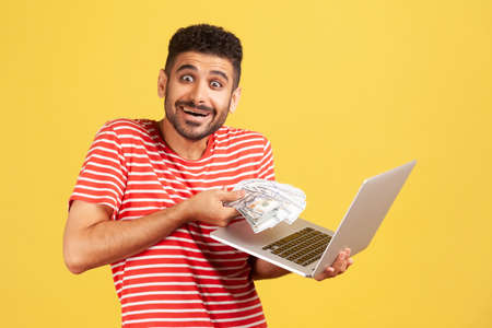 Happy cheerful man with beard in striped t-shirt holding laptop and lot of dollar bills, satisfied...