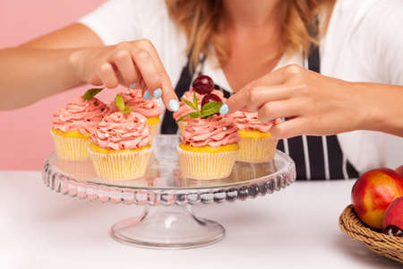 Close shot of many sweet cupcakes on grass plate, baker decorating sweet delicious muffins, homemade pastry getting ready for serving. Indoor studio shot isolated on pink background.の写真素材