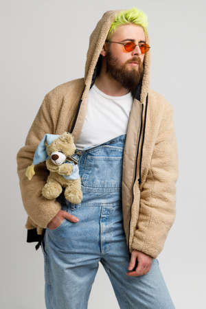 Attractive bearded man model wearing fashionable closing, has Teddy bear as accessory, keeping hand in pocket, looks away with thoughtful expression. Indoor studio shot isolated over gray background.の写真素材