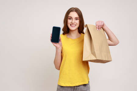 Smiling positive teenager girl in casual yellow T-shirt holding blank display smartphone and eco...