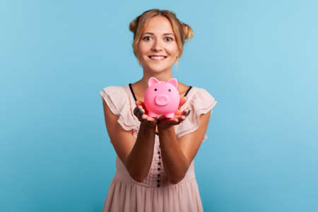 Attractive smiling young adult woman with pleasant appearance and blonde hair, showing piggy bank to camera, happy with money savings. Indoor studio shot isolated on blue background.の写真素材
