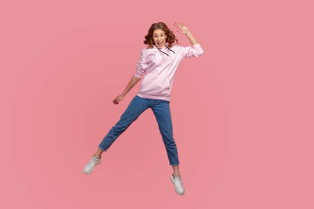 Full length portrait of cheerful young female in hoodie and jeans with toothy smile waving hand in...