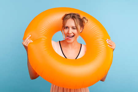 Funny female with two hair buns in dress holding orange rubber ring, having rest on resort, looking at camera and showing tongue out. Indoor studio shot isolated on blue background.の写真素材