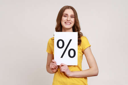 Smiling smart girl with brown wavy hair wearing yellow casual T-shirt holding paper and shows with...