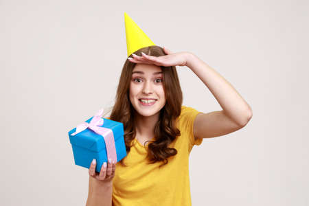 Portrait of teenager girl in T-shirt and party cone, holding gift box, looks far away with hand over eyes, waits future holidays, birthday celebration. Indoor studio shot isolated on gray background.の写真素材