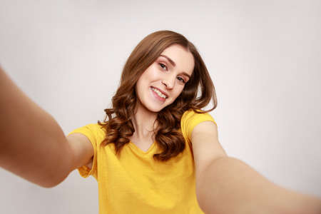 Portrait of beautiful teenager girl with brown hair in casual style T-shirt taking selfie, looking...