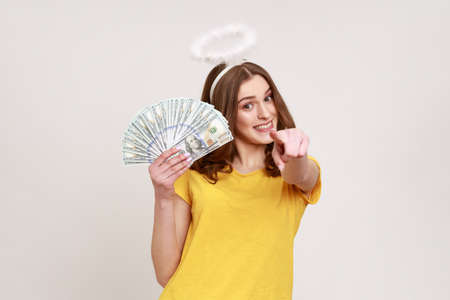Portrait of lottery winner lucky angelic girl with halo over head showing fan of money, holding dollar bills and pointing to camera with index finger. Indoor studio shot isolated on gray background.の写真素材