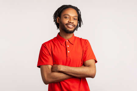 Young adult bearded man with dreadlocks wearing red casual T-shirt posing with confident facial expression and crossed arms, looking smiling at camera. Indoor studio shot isolated on gray background.の写真素材
