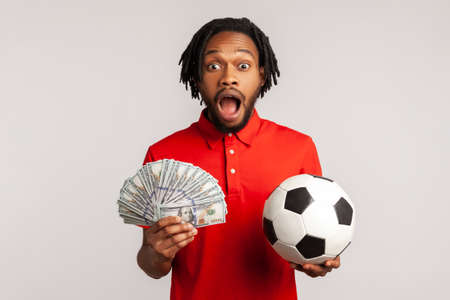 Yelling man with dreadlocks wearing red casual style T-shirt, holding soccer ball and hundred dollar banknotes, looking camera, betting and winning. Indoor studio shot isolated on gray background.の写真素材