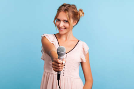 Young adult beautiful woman reporter in summer dress holding microphone, making interview and asking opinion, discussing important topics. Indoor studio shot isolated on blue background.の写真素材