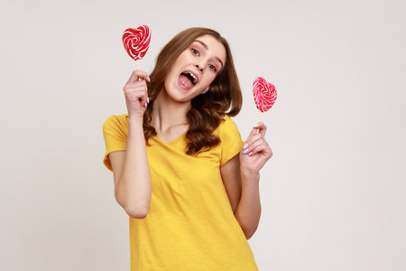 Portrait of cheerful playful woman in yellow casual T-shirt holding sweet round candy lollipop and looking at camera with excited childish expression. Indoor studio shot isolated on gray background.の写真素材