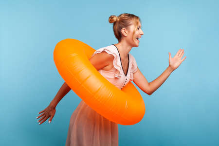 Side view of blonde woman with hair buns in summer dress running to sea, holding rubber ring, being hurry to relax in sea, expressing happiness. Indoor studio shot isolated on blue background.の写真素材